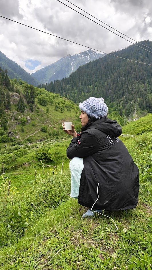 Varuna sitting in a Kashmir mountain meadow holding a cup, surrounded by green hills and pine trees